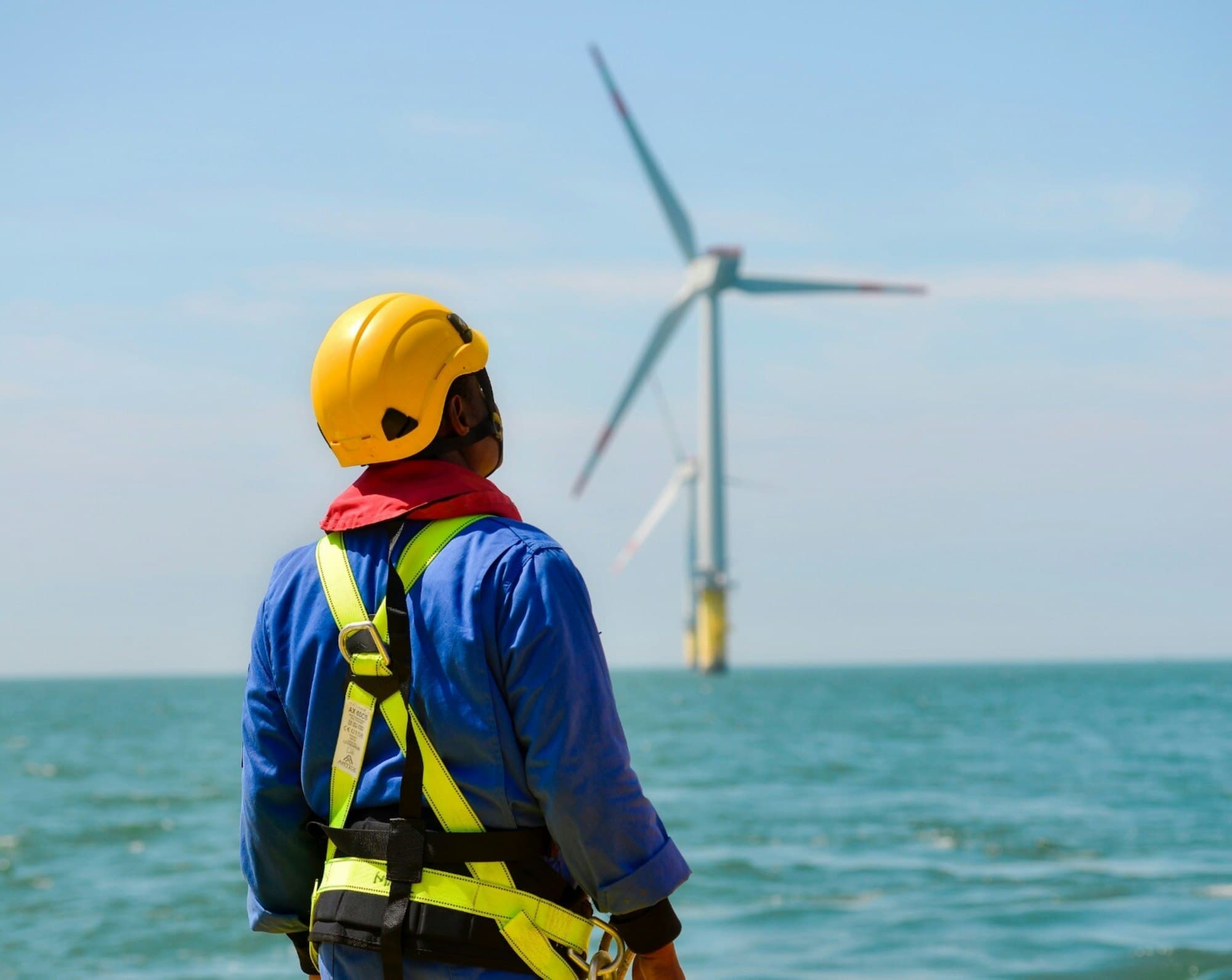 A worker in a hard hat and safety harness looking out at an offshore wind turbine