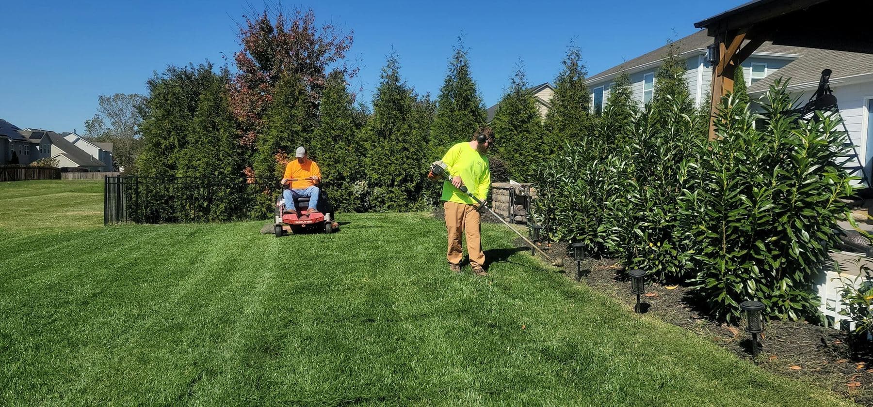 Two grounds crew members mowing and trimming along a hedge at a property