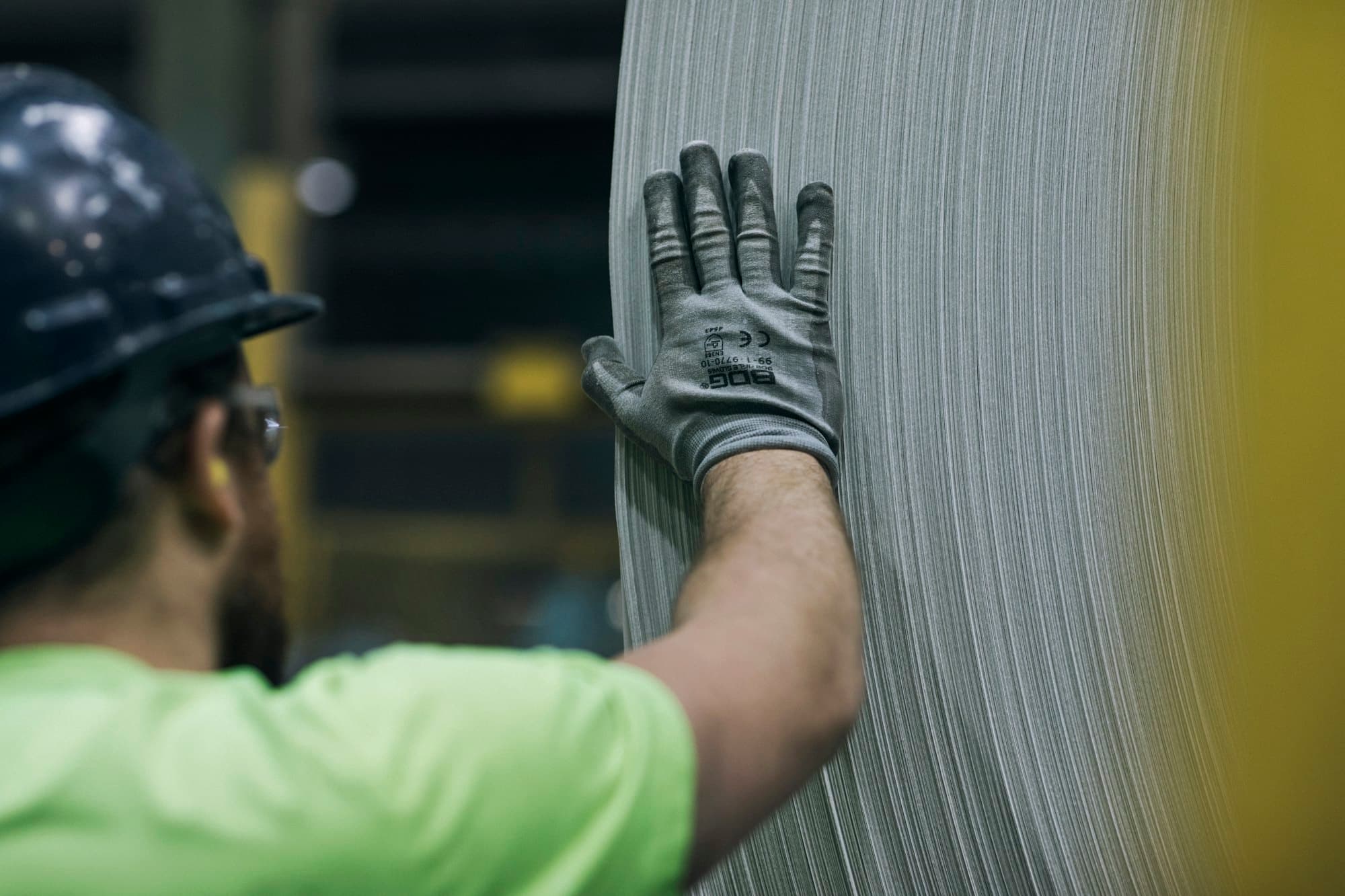 A worker in a hard hat and glove resting a hand on a large industrial roll