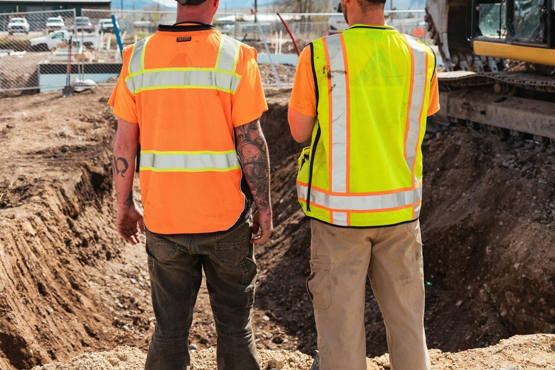 Two workers in high-visibility vests and hard hats looking into an excavation with an excavator behind them