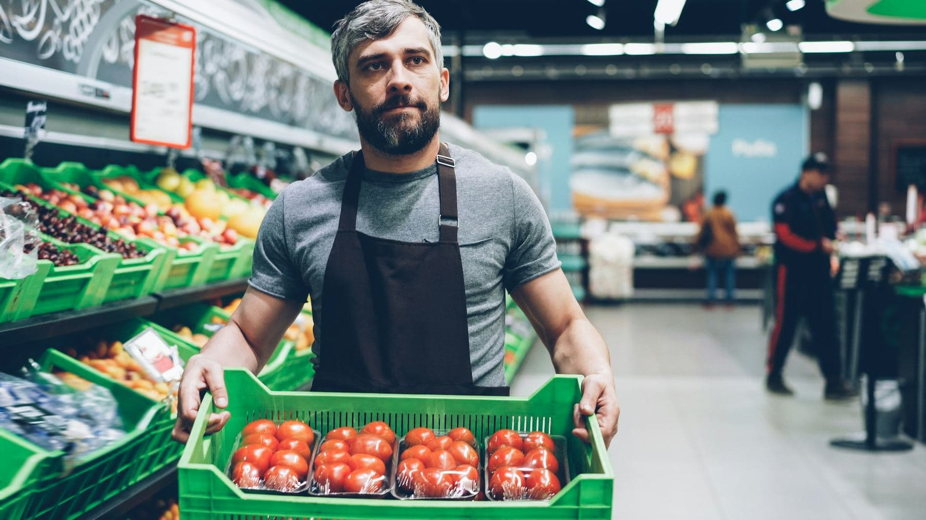 A grocery worker in an apron holding a crate of tomatoes in the produce aisle