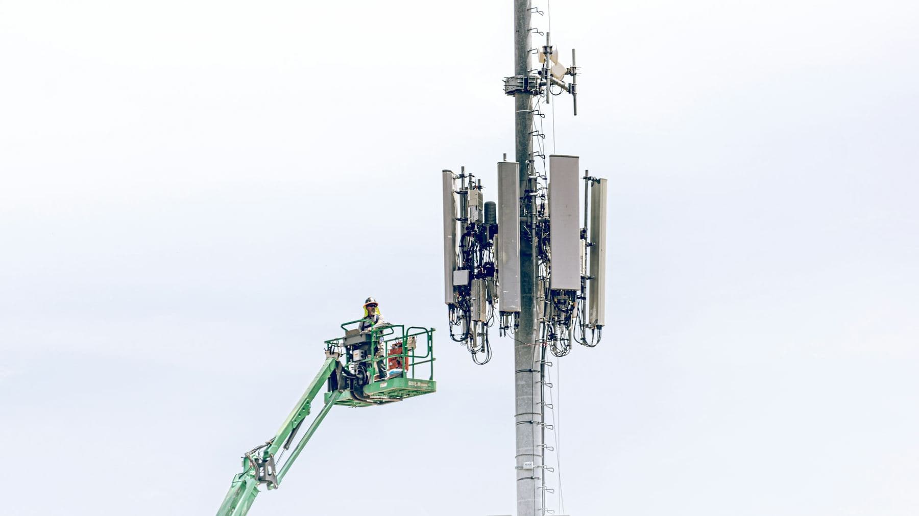 A technician in a lift basket working beside a cell tower against a pale sky