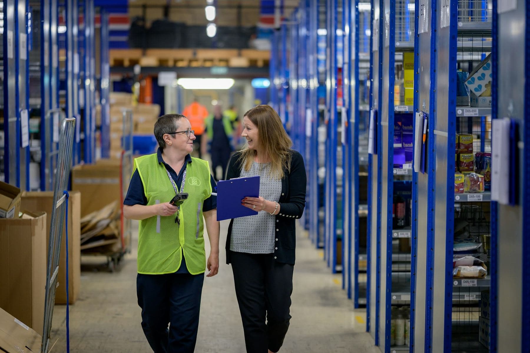 Two warehouse workers in high-visibility vests, one with a scanner and one with a clipboard, walking a racked aisle