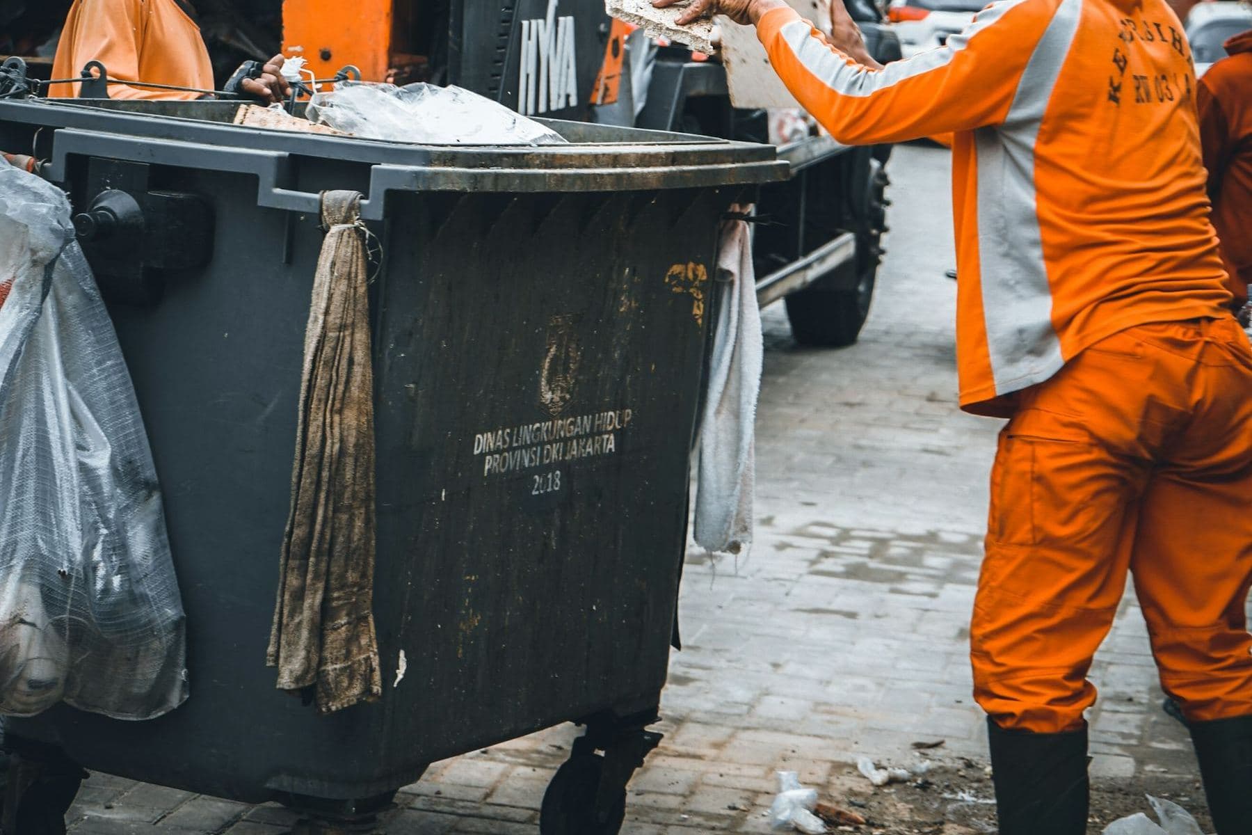 Sanitation workers in orange gear loading a collection truck beside a large bin